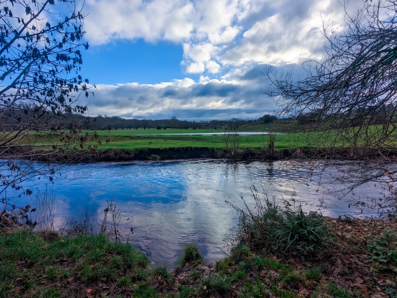 A river below fields and houses on a hill, bare trees and heavy clouds